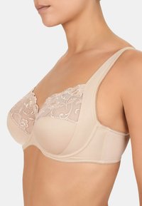 Soutien-gorge beige avec overlay en dentelle mettant en avant une broderie florale, des bonnets moulés, des bretelles larges et une fermeture par agrafes, offrant un design structuré.