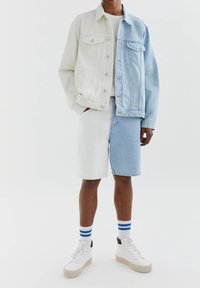 Veste en jean à design color-block avec une moitié bleu clair et une moitié bleu foncé, associée à un short en jean blanc et bleu. Baskets blanches avec des accents bleus.