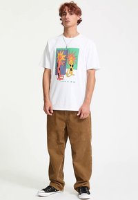 White graphic t-shirt featuring colorful cartoon designs of figures, paired with brown corduroy pants and black sneakers with white soles.