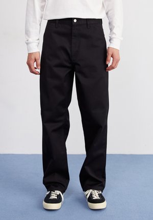 Person trägt schwarze Hosen mit geradem Bein, weißes Langarmoberteil und schwarze Sneakers mit weißen Schnürsenkeln steht auf blauem Boden.