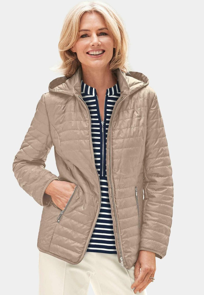 Beige gesteppte Jacke mit Kapuze, mit Reißverschluss vorne und seitlichen Taschen. Getragen über einem gestreiften Oberteil in Marineblau und Weiß.