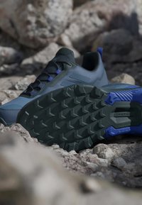 Grå sportskor med en strukturerad yta, prydda med svarta snören och blå accenter. Sulan har ett robust mönster för bra grepp.