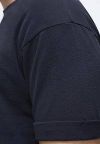 Gros plan d'une personne portant un t-shirt uni bleu marine foncé à manches courtes avec un revers plié sur la manche.