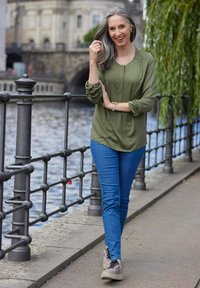 Olivgrüne Bluse, lange Ärmel, weicher Stoff; blaue Skinny-Jeans; hellfarbige Sneakers mit schwarzen Schnürsenkeln; Wasserfront-Hintergrund mit Pflanzen.