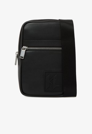 Must ristmikuga kandekott, millel on hõbedane tõmblukuga tasku, paks must õlarihm ja esiküljel reljeefse logomärgiga patch.