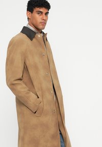 Tan coat with a smooth texture, snap buttons, and a dark collar. Features side pockets and a straight cut for a modern look.