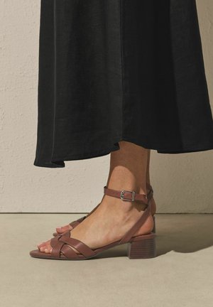 Fod iført brun læder sandal med ankelrem og lav blokhæl, kombineret med en lang sort nederdel mod en beige væg og gulv.