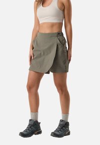 Femme portant un crop top beige, une jupe portefeuille olive avec des poches, des bottes de randonnée grises et des chaussettes gris clair, debout devant un fond blanc.