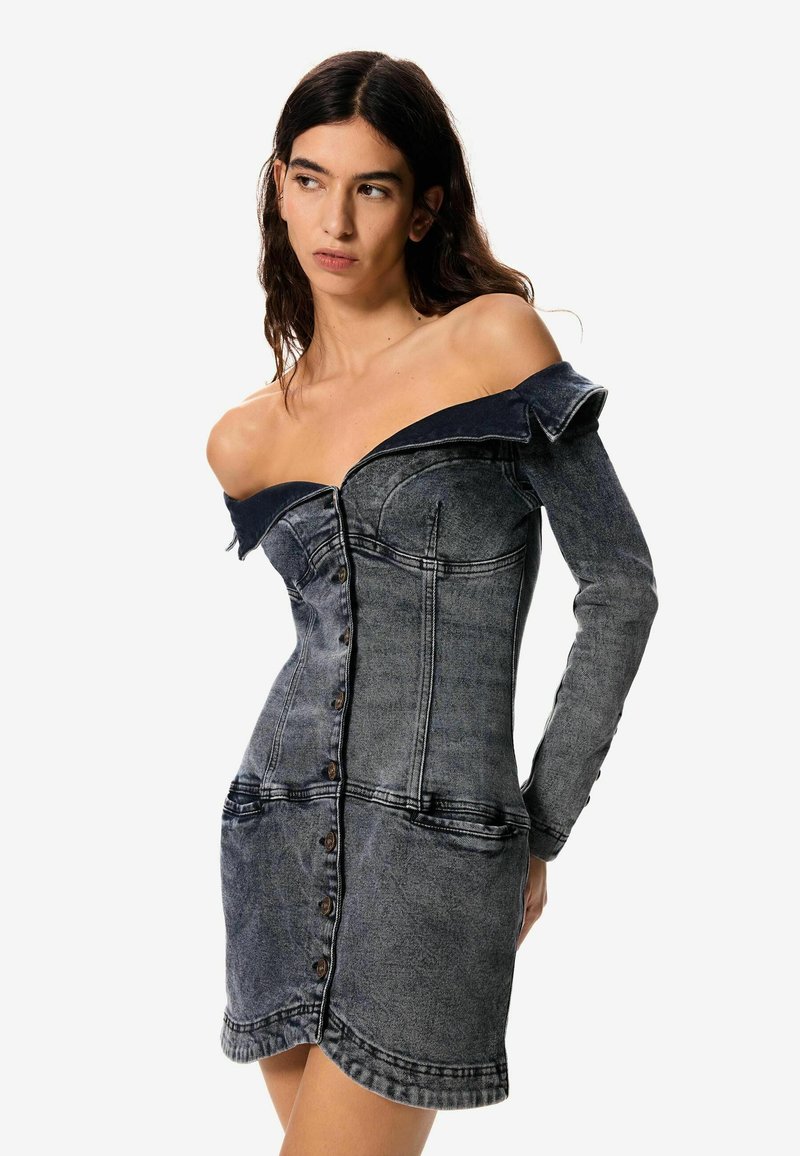 Žena u izblijedjeloj crnoj traper haljini bez ramena s dugim rukavima i kopčanjem, gledajući ustranu na neutralnoj svijetloj pozadini.