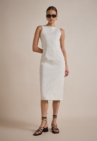 Weißes, knielanges, ärmelloses Kleid mit geradem Schnitt und Nahtdetail an der Vorderseite. Getragen mit schwarzen Riemchensandalen und Sonnenbrille.