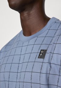 Lys blå t-shirt med et gittermønster og sort tekst, der siger "spurs." Har et rektangulært logo-patch på brystet. Blød stofstruktur.