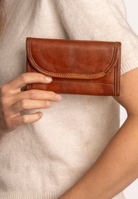 Brown leather wallet with a curved flap, visible stitching, and a compact rectangular shape. Held in a hand against a light sweater background.
