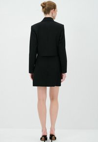 Abito blazer su misura nero con design cropped, maniche lunghe e dettagli con bottoni. Tessuto liscio, silhouette aderente, lunghezza al ginocchio.