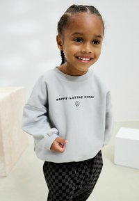 Svetlosivý sveter s nápisom "ŠŤASTNÝ MALÝ ČLOVEK" a smajlíkom. Má voľný strih, rebrované manžety a kombinuje sa s čiernymi kockovanými nohavicami.