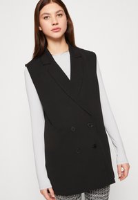 Chaleco negro sin mangas con un frente cruzado, que presenta botones prominentes y solapas marcadas. Tela suave y silueta estructurada.