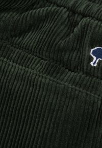 Donkergroene corduroy stof met ribbeltextuur en een klein geborduurd blauw en wit boomlogo rechtsboven.