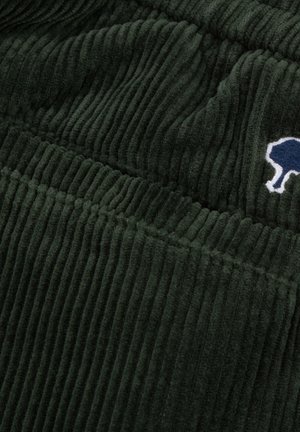Tessuto in velluto a coste verde scuro con texture a costine e un piccolo logo di un albero ricamato in blu e bianco nell'angolo in alto a destra.