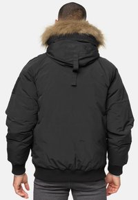 Sort dunjakke med hætte pyntet med pelskant, ribbede manchetter og bund, samt en tydeligt polstret design. Har en fastgørelsesrem på kraven bagpå og en sidelap.