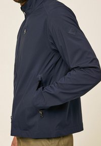 Homme portant une veste zippée bleu marine avec des poches zippées et un petit écusson logo sur la manche supérieure, main dans la poche latérale.