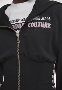 Čierna zipová mikina s textúrovanou látkou, na ktorej sa nachádza vyšívané logo "Versace Jeans Couture" v ružovej a bielej farbe. Postranné detaily s čipkou.