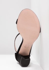 Sandale à talon bas en daim noir reposant sur une surface blanche, montrant la semelle lisse couleur beige avec motifs antidérapants et texte de la marque.