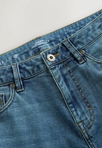 Gros plan de la ceinture d'un jean en denim bleu slim fit avec un bouton métallique, des passants de ceinture, des coutures et une étiquette imprimée indiquant "NEXT PERFORMANCE SLIM FIT."