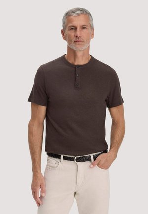 Man met grijs haar en baard, gekleed in een donkerbruin henley-shirt met korte mouwen, ingestopt in gebroken witte broek met een zwarte gevlochten riem.