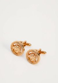 Ζευγάρι στρογγυλών μανικετόκουμπων σε χρυσό τόνο, χαραγμένα με κεφάλι Μέδουσας και μοτίβο ελληνικού κλειδιού, τοποθετημένα σε λευκή επιφάνεια.