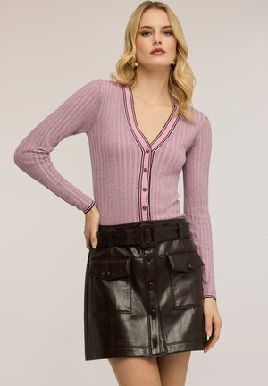 Mujer con un cárdigan rosa acanalado con botones y ribete negro, y una falda de cuero negra de talle alto con bolsillos delanteros y cinturón.