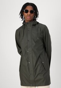 Groene waterdichte jas met een capuchon, drukknoopsluiting en zijzakken. Soepele textuur en een relaxte pasvorm. Draagt een ronde zonnebril.