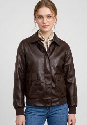 Młoda kobieta nosząca przejrzyste okulary, jasny wzorzysty szalik, ciemnobrązową skórzaną kurtkę i niebieskie jeansy, stojąca na jednolitym tle.