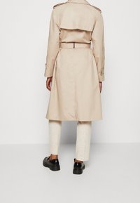 Béžový trenčkot s páskem v pase, dvojitými ramenními epolety a rozparkem na zádech. Kombinováno s krémovými kalhotami a černými botami.