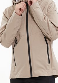 Veste légère beige avec une fermeture éclair à l'avant, deux poches latérales et une coupe ajustée. Équipée de caractéristiques réglables et d'une texture douce.