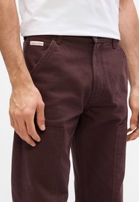 Pantaloni in denim borgogna con tasche anteriori e etichetta Calvin Klein. Il tessuto ha una texture liscia, una vestibilità comoda e cuciture robuste.