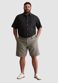 Schwarzes Leinenhemd mit kurzen Ärmeln, einer Brusttasche und weißen Knöpfen, kombiniert mit grau gestreiften Shorts und weißen Sneakers mit beigen Sohlen.