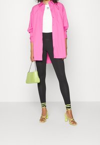 Chemise rose surdimensionnée, crop top blanc, leggings noirs, talons jaunes à lanières avec des accents, et un petit sac à main vert texturé.
