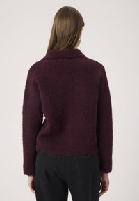 Una donna con capelli lunghi e lisci di colore castano indossa un maglione lungo a maniche lunghe di un viola scuro e dei pantaloni neri, in piedi contro uno sfondo neutro.