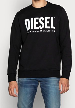 Homme portant un sweat-shirt noir avec le texte "DIESEL FOR SUCCESSFUL LIVING", assorti à un jean bleu sur fond uni.