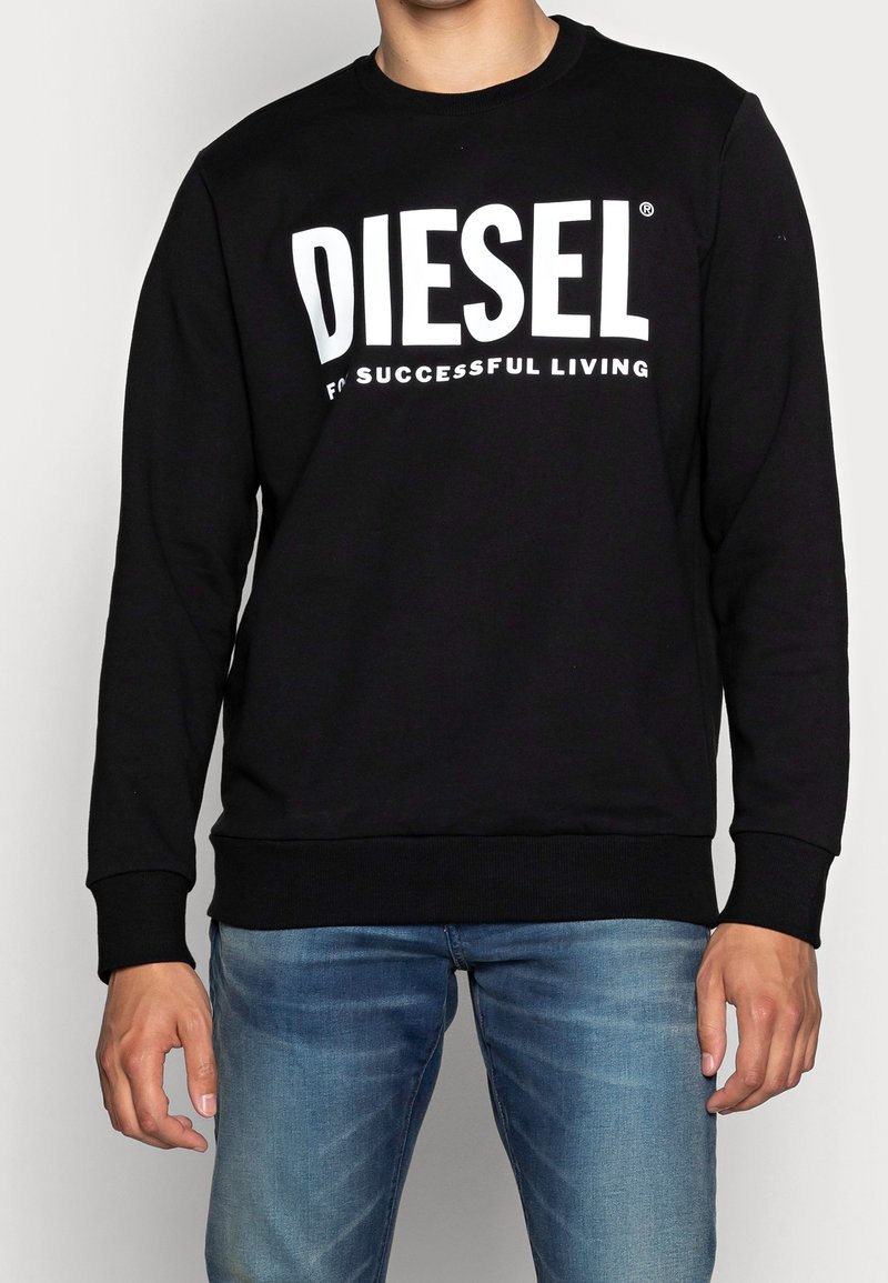 Homme portant un sweat-shirt noir avec le texte "DIESEL FOR SUCCESSFUL LIVING", assorti à un jean bleu sur fond uni.