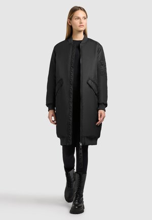 Black long bomber jacket with a smooth finish, zip closure, side pockets, ribbed cuffs, and textured patch on the sleeve. Paired with black leggings and combat boots.