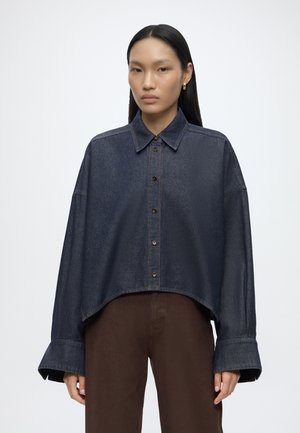 Frau mit langen schwarzen Haaren, die ein dunkelblaues oversized Jeanshemd und braune Hosen trägt, steht vor einem schlichten weißen Hintergrund.