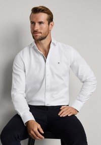 Chemise blanche à col pointu, ornée d'un logo brodé sur la poitrine et d'un détail de rayures contrastantes au niveau du col.