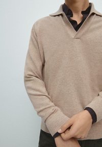Maglione beige lavorato a maglia con scollo a V e design con colletto. Presenta polsini a coste e una finitura testurizzata. Indossato sopra una camicia scura.
