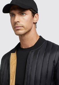 Black puffer jacket with vertical quilted pattern, featuring a gold lining, paired with a black cap and black crew neck shirt.
