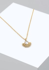 Collana in tono oro con pendente a forma di esplosione solare, caratterizzata da una pietra centrale trasparente e una catena testurizzata. Il design enfatizza le forme geometriche e la luce.