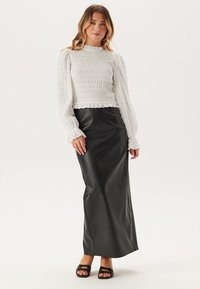 Bílý top s volánkovými detaily a dlouhým rukávem v kombinaci s elegantní dlouhou černou koženou sukní. Modelka má na sobě otevřené černé sandály.