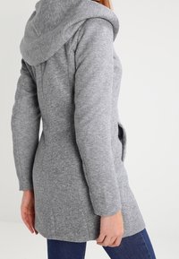 Vrouw in een aansluitende grijze hoodie met lange mouwen, staande met haar rug iets naar links gekeerd, tegen een eenvoudige lichte achtergrond.
