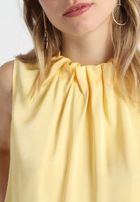 Top sin mangas amarillo con escote fruncido y textura de tela suave. Destacados pendientes de aro dorados que complementan el diseño.