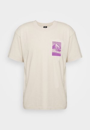 Beżowy T-shirt z krótkim rękawem i okrągłym dekoltem oraz małą fioletową grafiką góry na lewej części klatki piersiowej.
