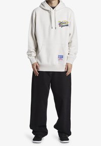 Jasnobeżowa bluza z kapturem, z kieszenią typu kangurka, sznurkiem w kapturze i wielokolorowym logo na piersi. Połączona z luźnymi czarnymi spodniami.